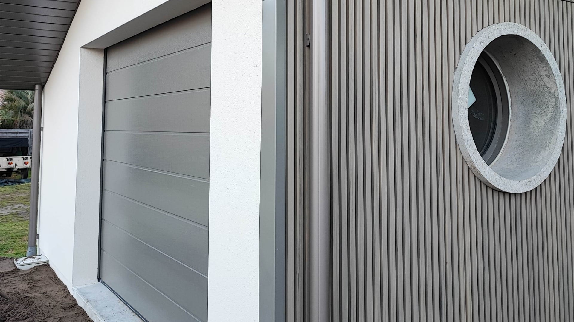 Construction maison Vieux-Boucau : Villa de 174 m² avec 3 suites - Maison Landes Habitat Porte de garage grise avec un mur en panneaux verticaux et une fenêtre circulaire.